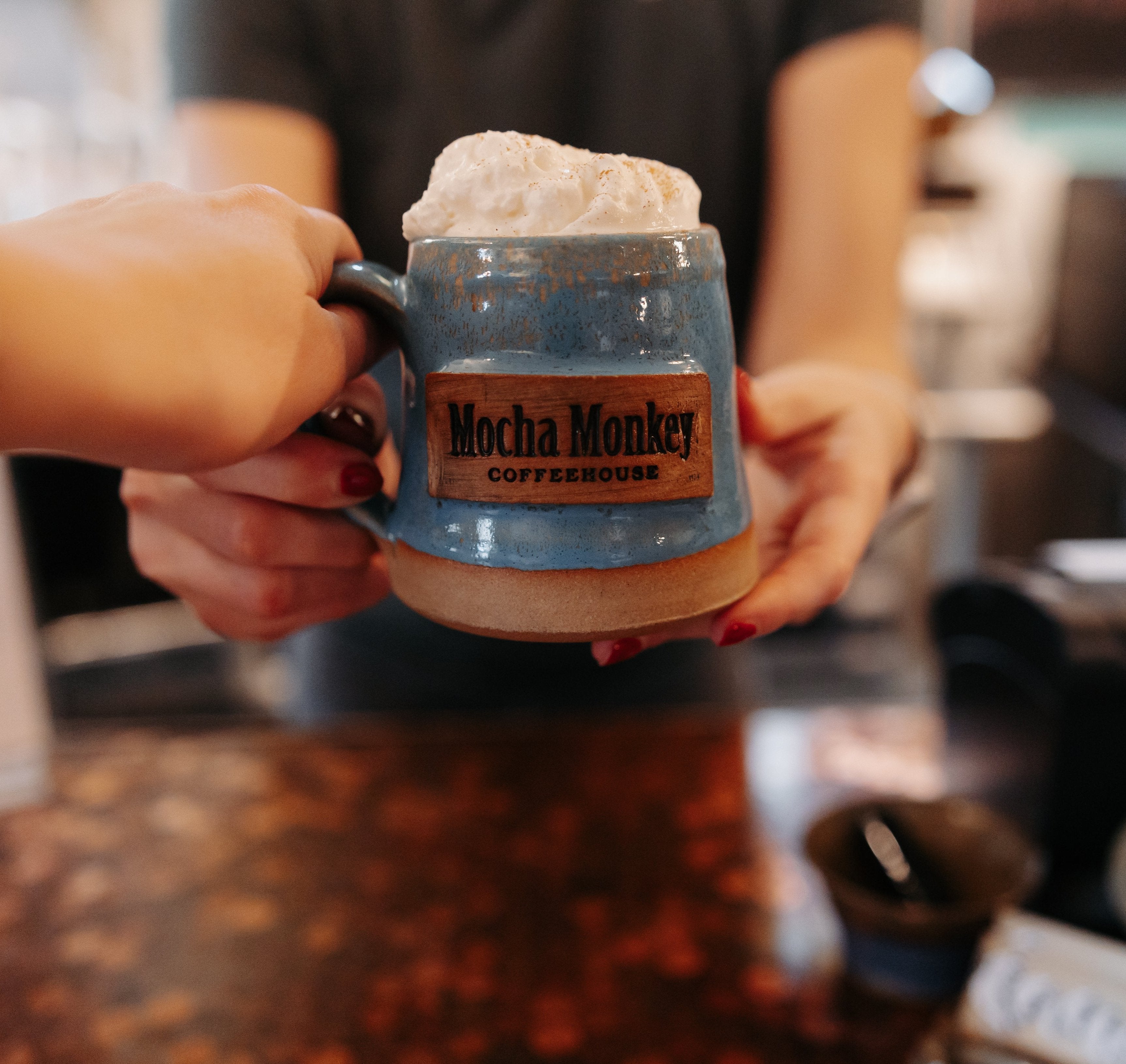 Barista handing a person a coffee with whipped cream in a handmade blue pottery mug branded with Mocha Monkey Coffeehouse