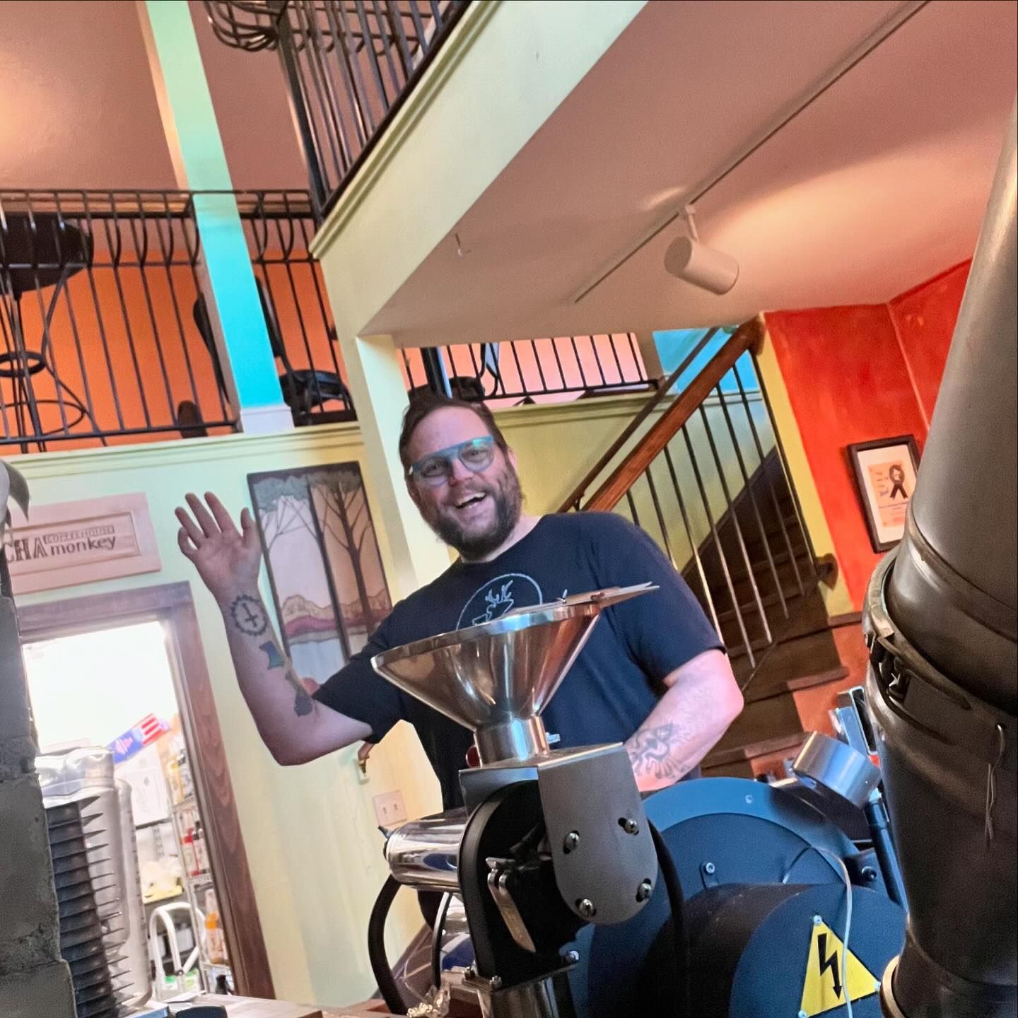 A smiling John Pennucci waving from behind Marcel the Coffee Roaster at the Mocha Monkey Downtown Waconia Coffeehouse