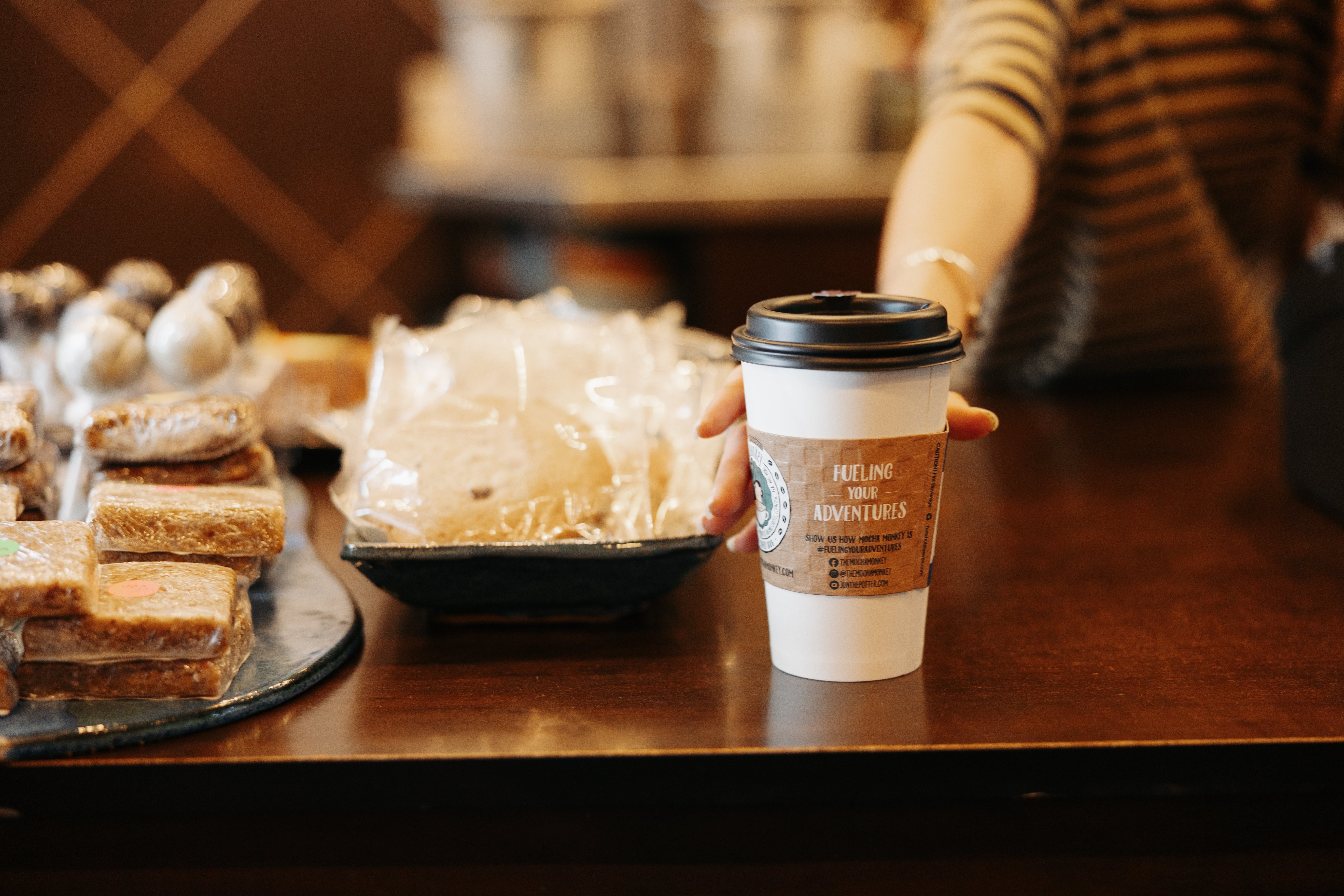 A person reaching for their coffee on a counter filled with baked goods