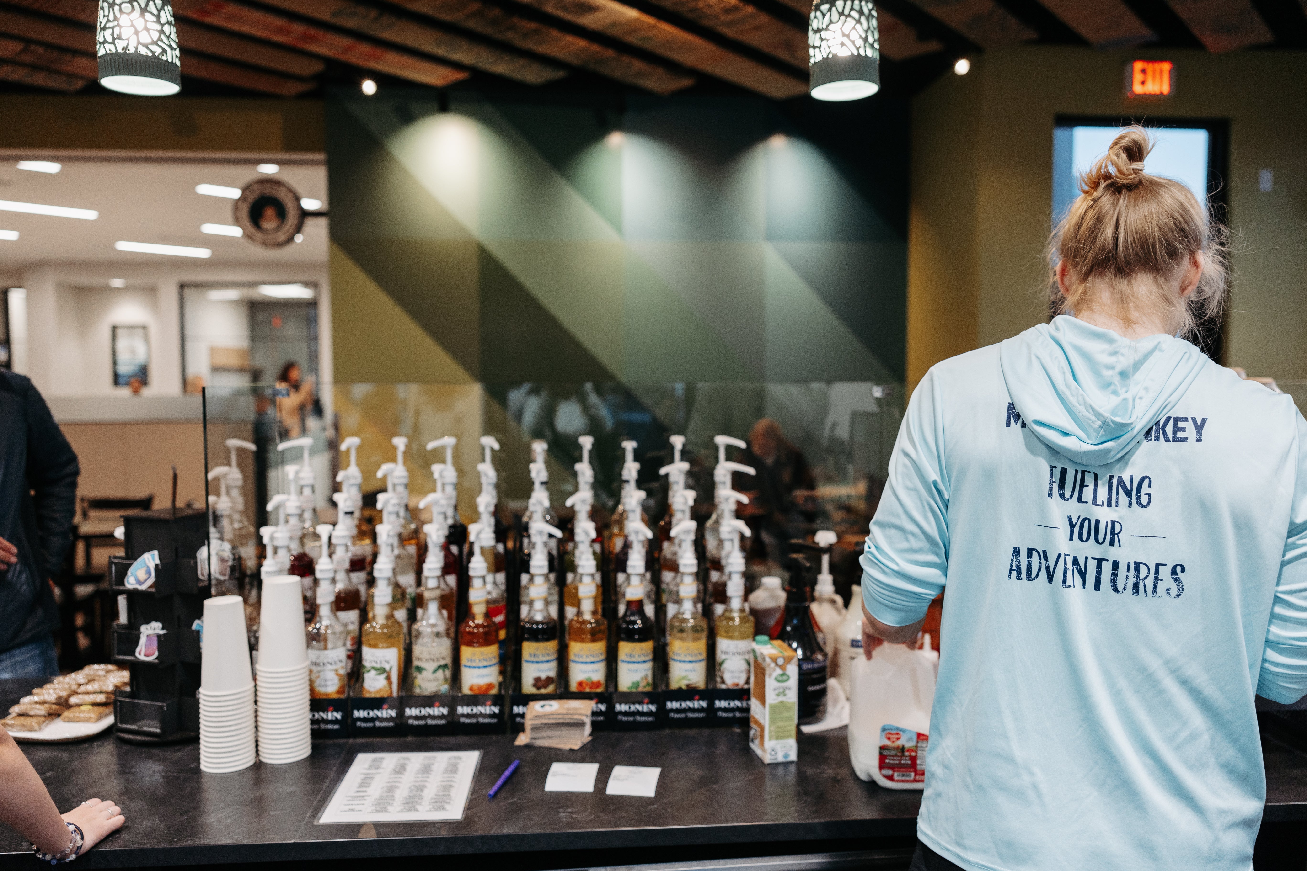 The back of barista wearing a Mocha Monkey Fueling Your Adventures hoodie with bottles of flavoring in the foreground